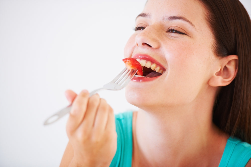 A young woman with long hair smiles as she takes a bite of a strawberry on a fork, set against a plain white background.