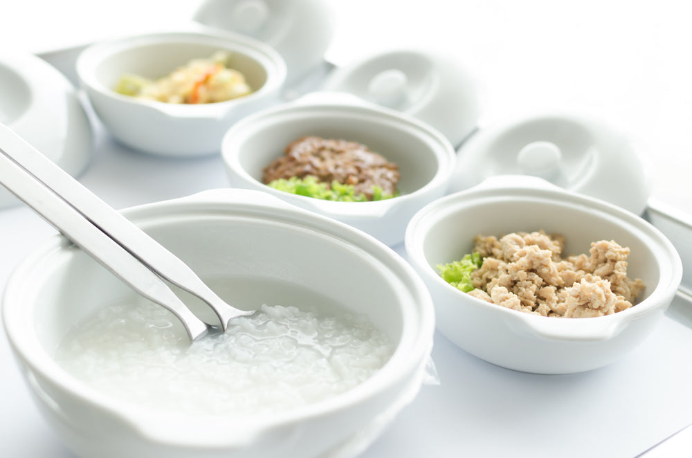 White bowls with rice porridge and small portions of minced meat and vegetables arranged on a bright table.