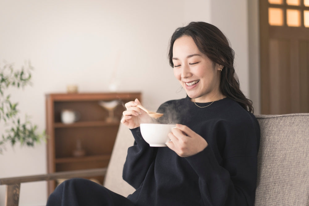 Woman sitting on a couch smiling while holding and eating from a steaming bowl of food.