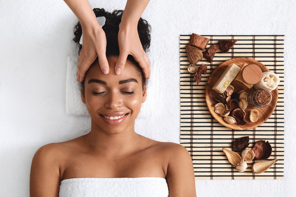 Woman smiling with eyes closed while receiving a forehead massage beside natural spa items.