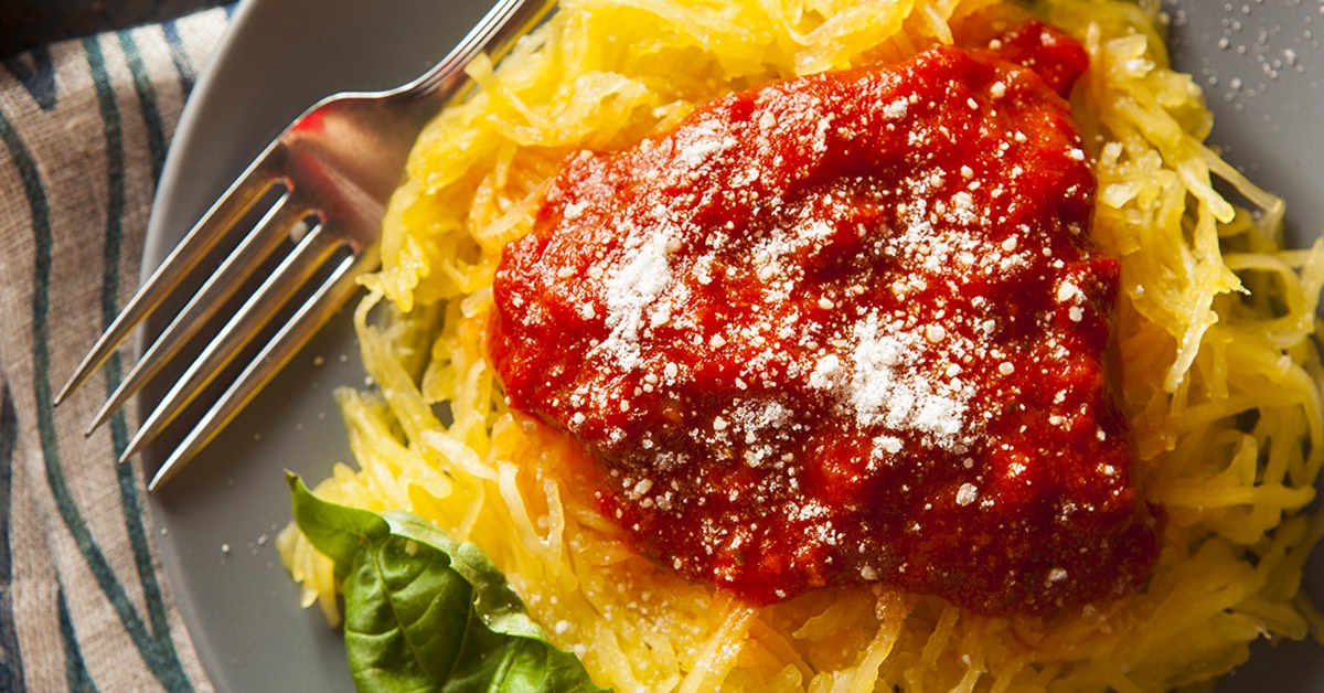 A plate of spaghetti squash topped with rich red marinara sauce and sprinkled with grated cheese, accompanied by a fork and a fresh basil leaf, sits on a textured tablecloth.
