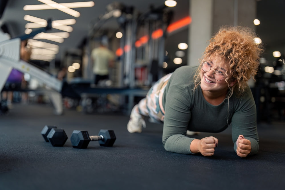 A person with curly hair and glasses smiles while doing a plank exercise on a gym floor, with dumbbells nearby and other gym-goers visible in the background.