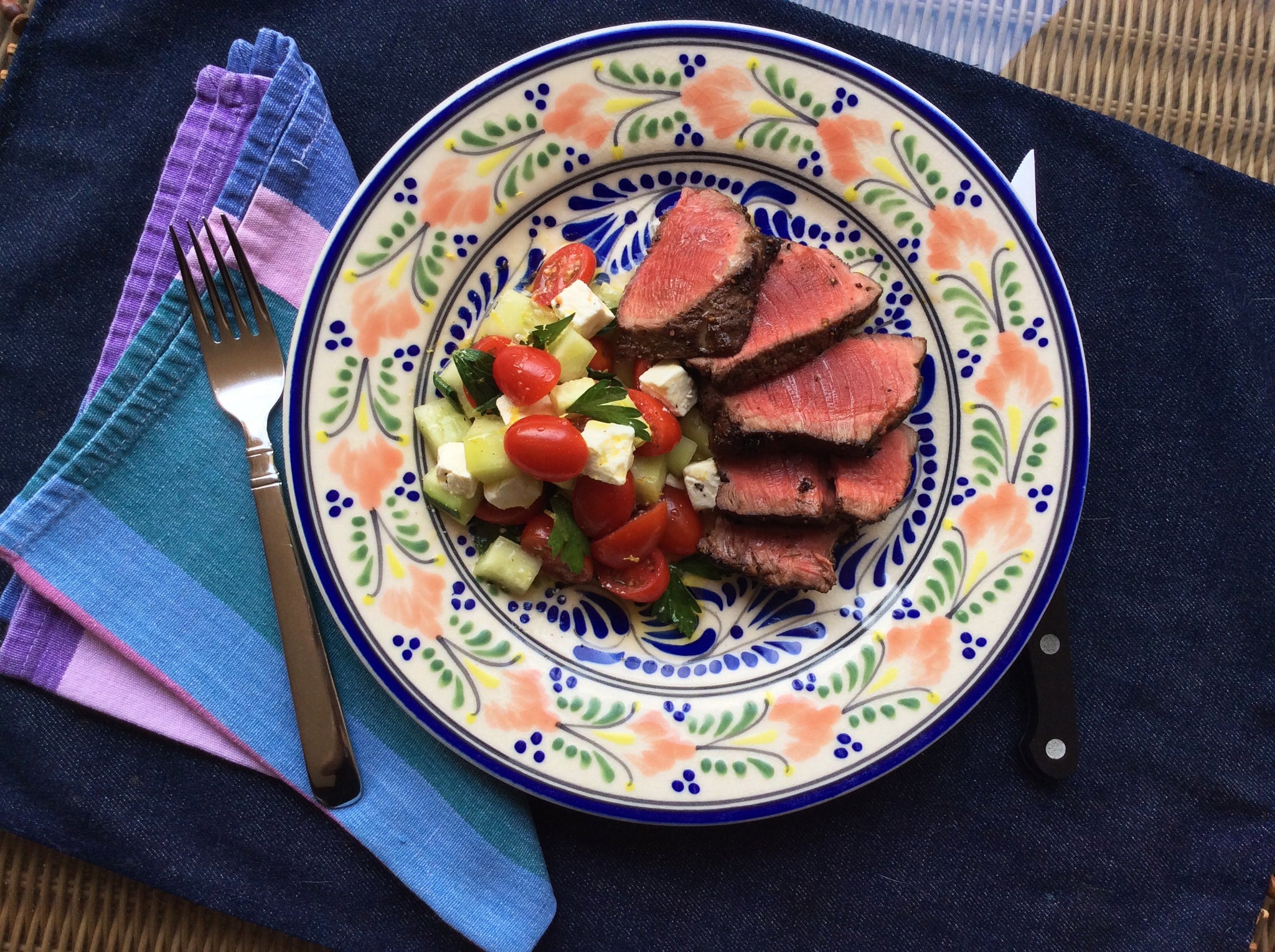 Sliced seared beef with a tomato, cucumber, and feta salad on a colorful patterned plate.