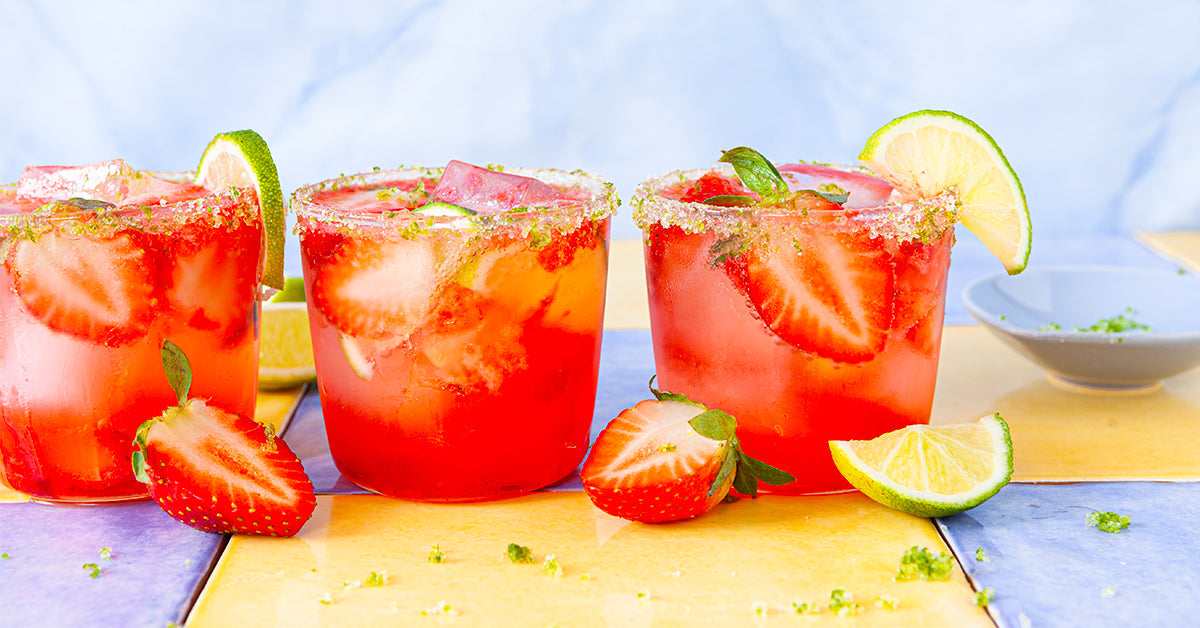 Three colorful cocktails with strawberries and lime slices sit on a bright, patterned table, garnished with mint leaves and a sugared rim, alongside fresh fruit and a small bowl of lime zest.