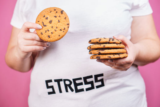 A person in a white shirt with the word "STRESS" printed on it holds a chocolate chip cookie in one hand and a stack of four cookies in the other, set against a pink background.
