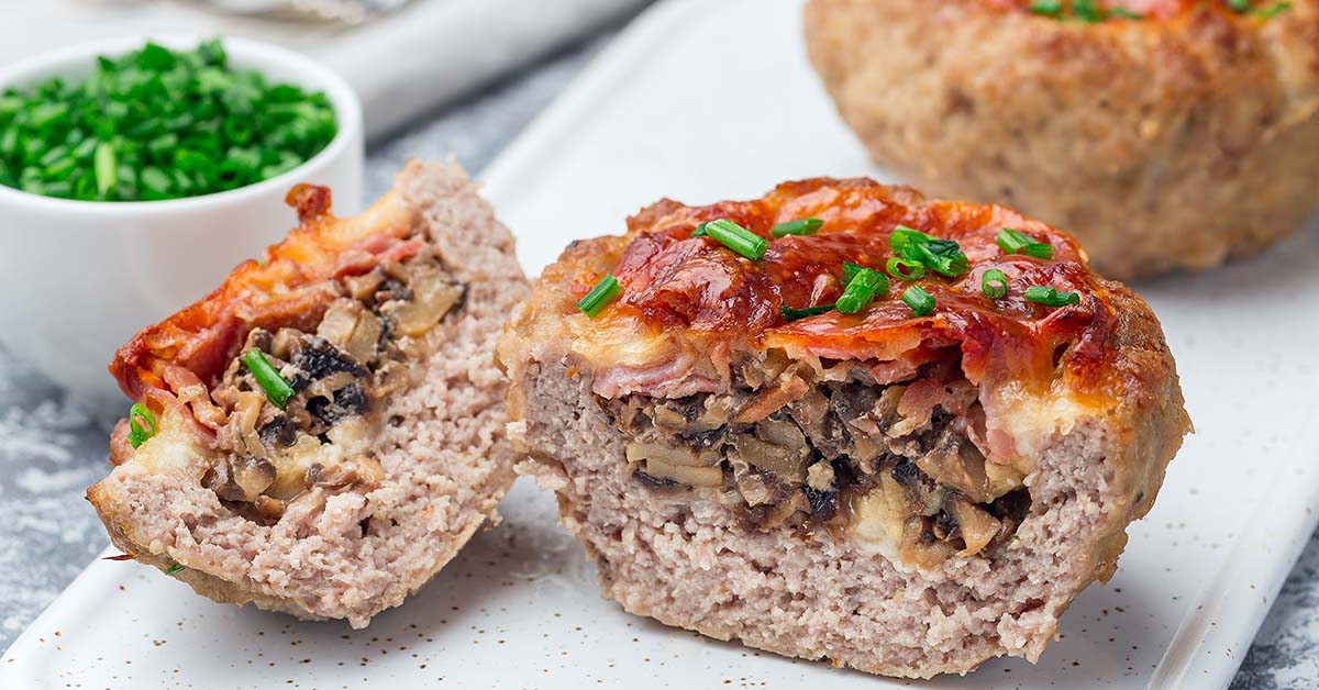 Stuffed meatloaf cups are displayed on a white plate, one cut in half to reveal a filling of mushrooms and cheese, topped with a glossy glaze and garnished with chopped chives, alongside a bowl of fresh chives.