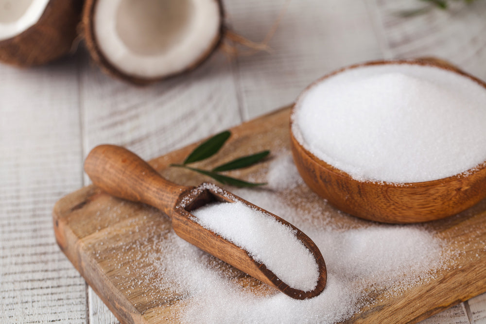 A wooden scoop filled with white sugar rests on a cutting board beside a bowl of sugar, with coconut halves and green leaves in the background on a light wooden surface.