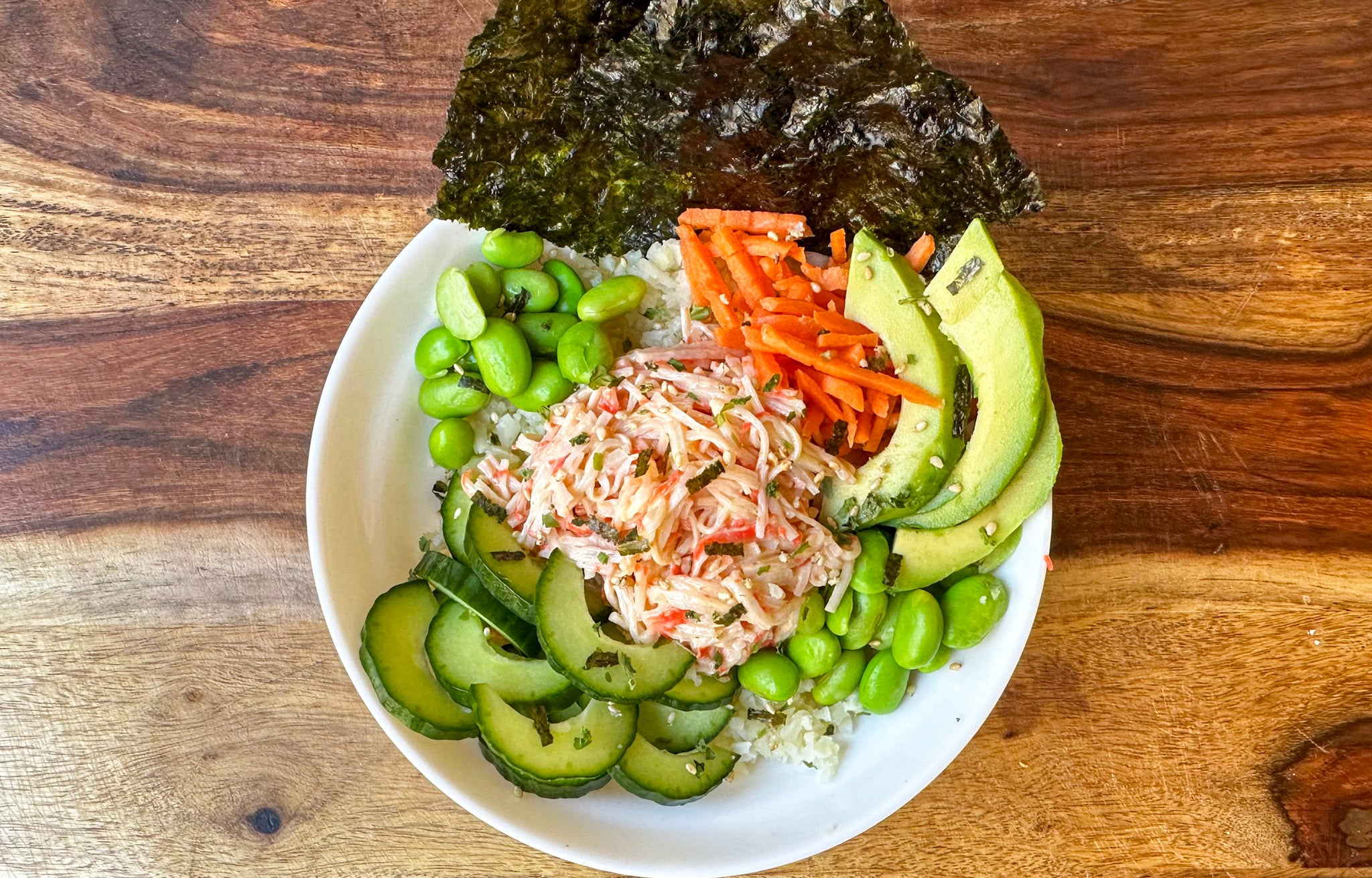 A white bowl filled with a lower carb sushi bowl features sliced cucumbers, avocado, shredded carrots, edamame, crab meat, and a sheet of nori, all arranged on a wooden table.