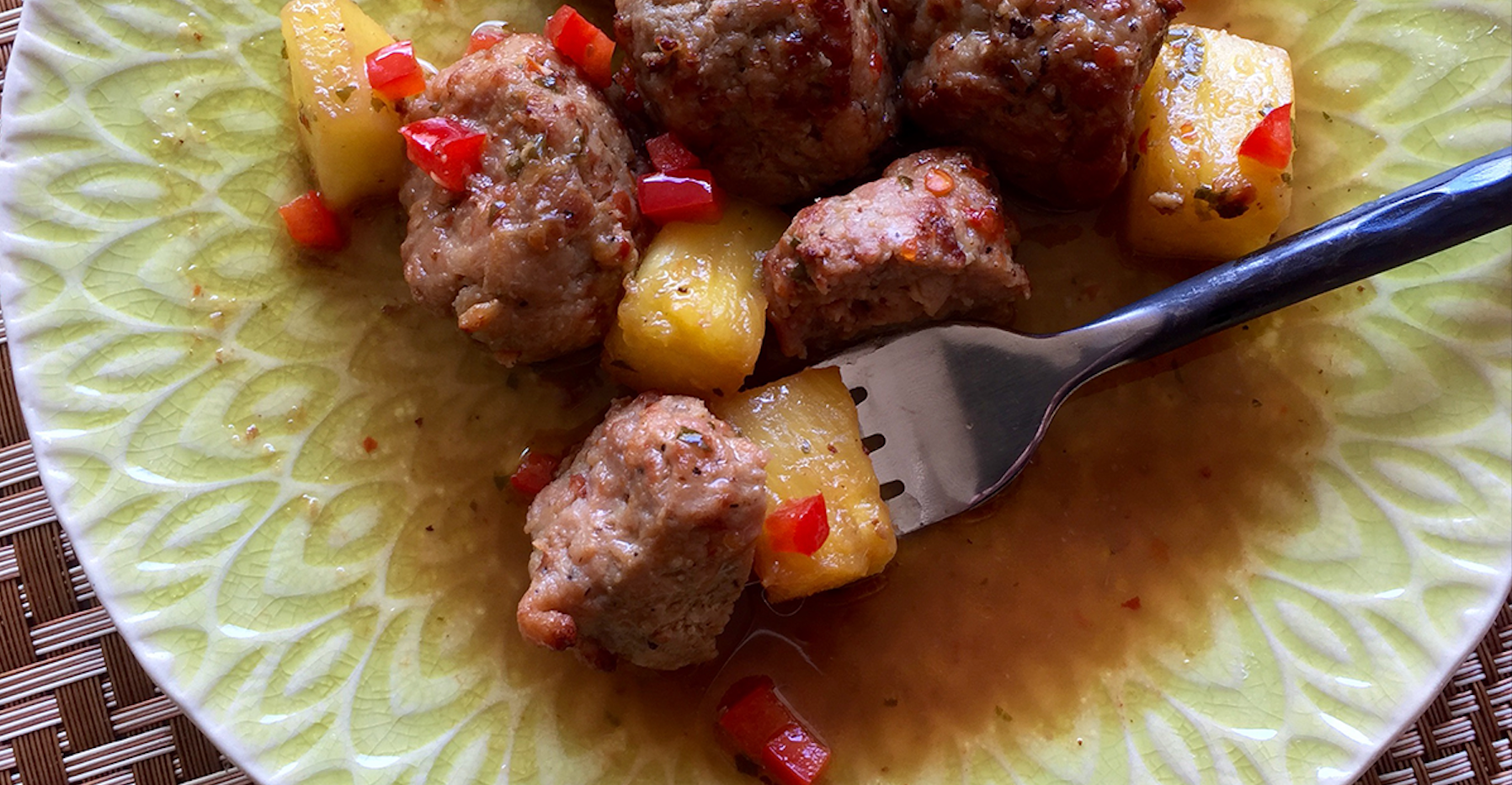 Meatballs and pineapple chunks with diced red peppers on a green plate, with a fork picking up a piece.