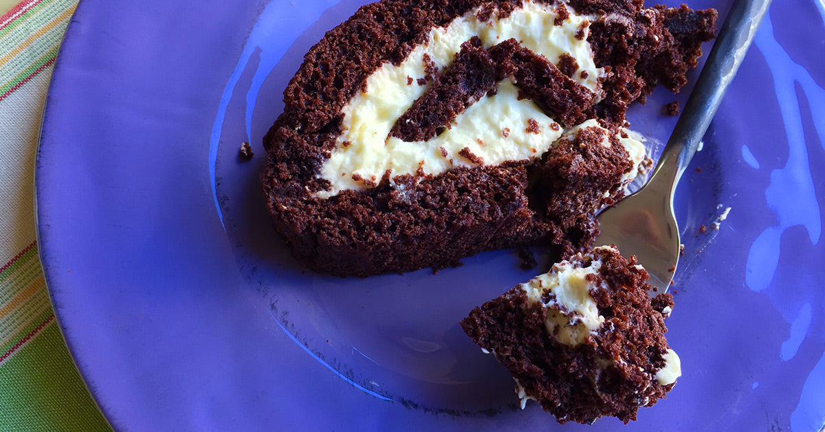 Slice of chocolate cake roll with cream filling on a purple plate, with a fork holding a bite.