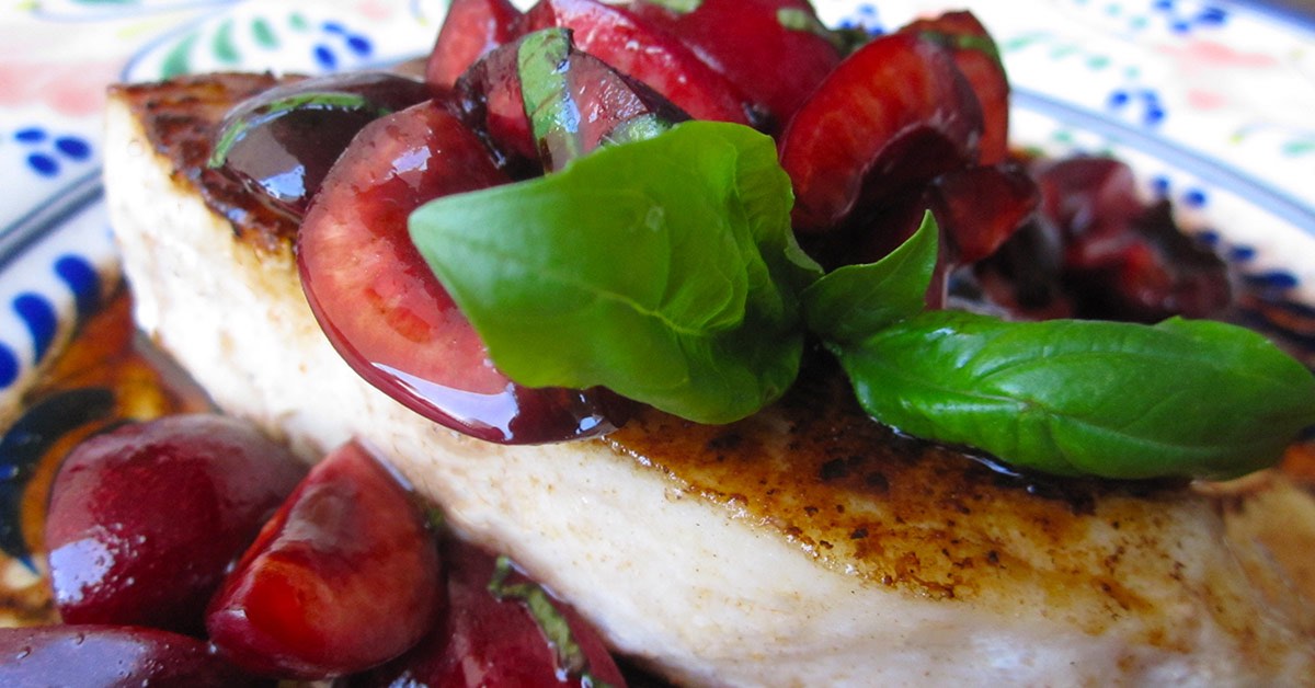 A piece of seared fish is topped with glossy, chopped cherries and fresh basil, presented on a colorful plate with decorative patterns.