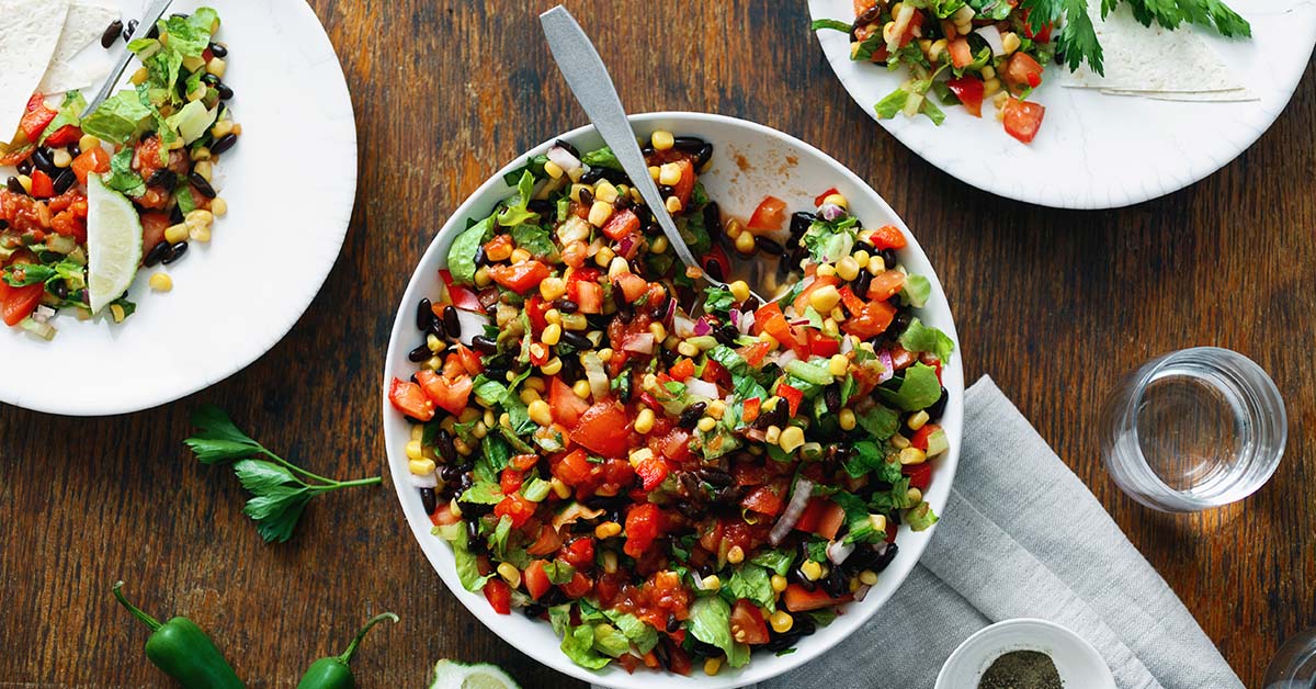 A vibrant bowl of Snappy Taco Salad, filled with lettuce, tomatoes, corn, and black beans, sits on a wooden table next to a plate with a small serving and a glass of water.