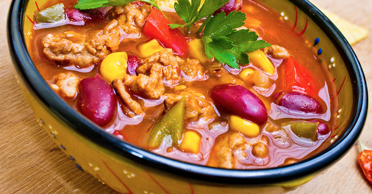 Close-up of a hearty soup with beans, corn, peppers, and meat in a decorative bowl.