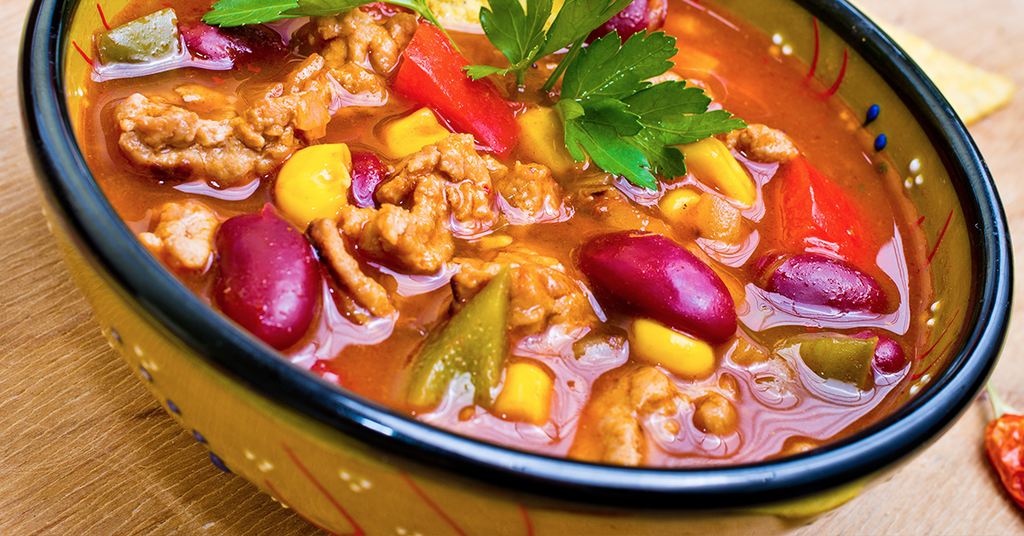 Close-up of a hearty soup with beans, corn, peppers, and meat in a decorative bowl.
