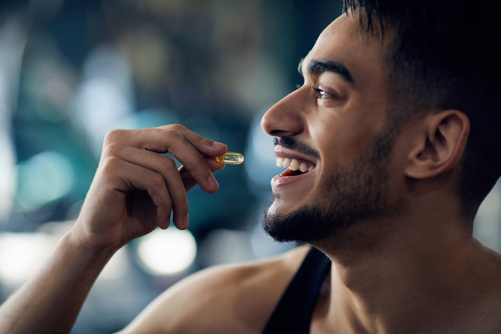 A smiling man holds a yellow capsule near his mouth, appearing happy and engaged, in a bright, blurred indoor setting that suggests a gym or fitness area.
