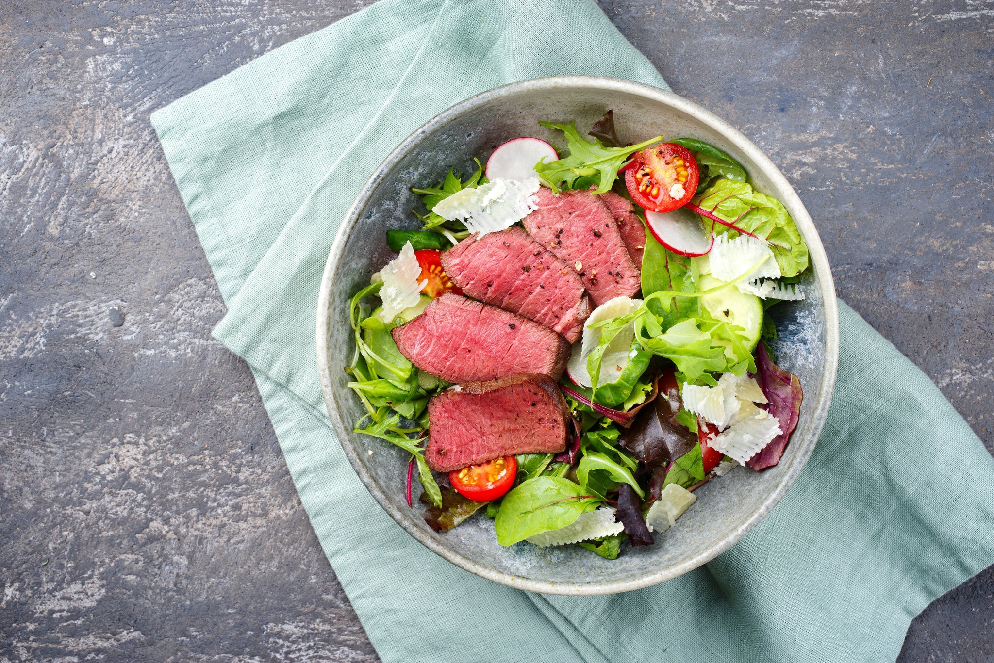 Bowl of mixed green salad topped with sliced medium-rare steak, tomatoes, radishes, and Parmesan shavings