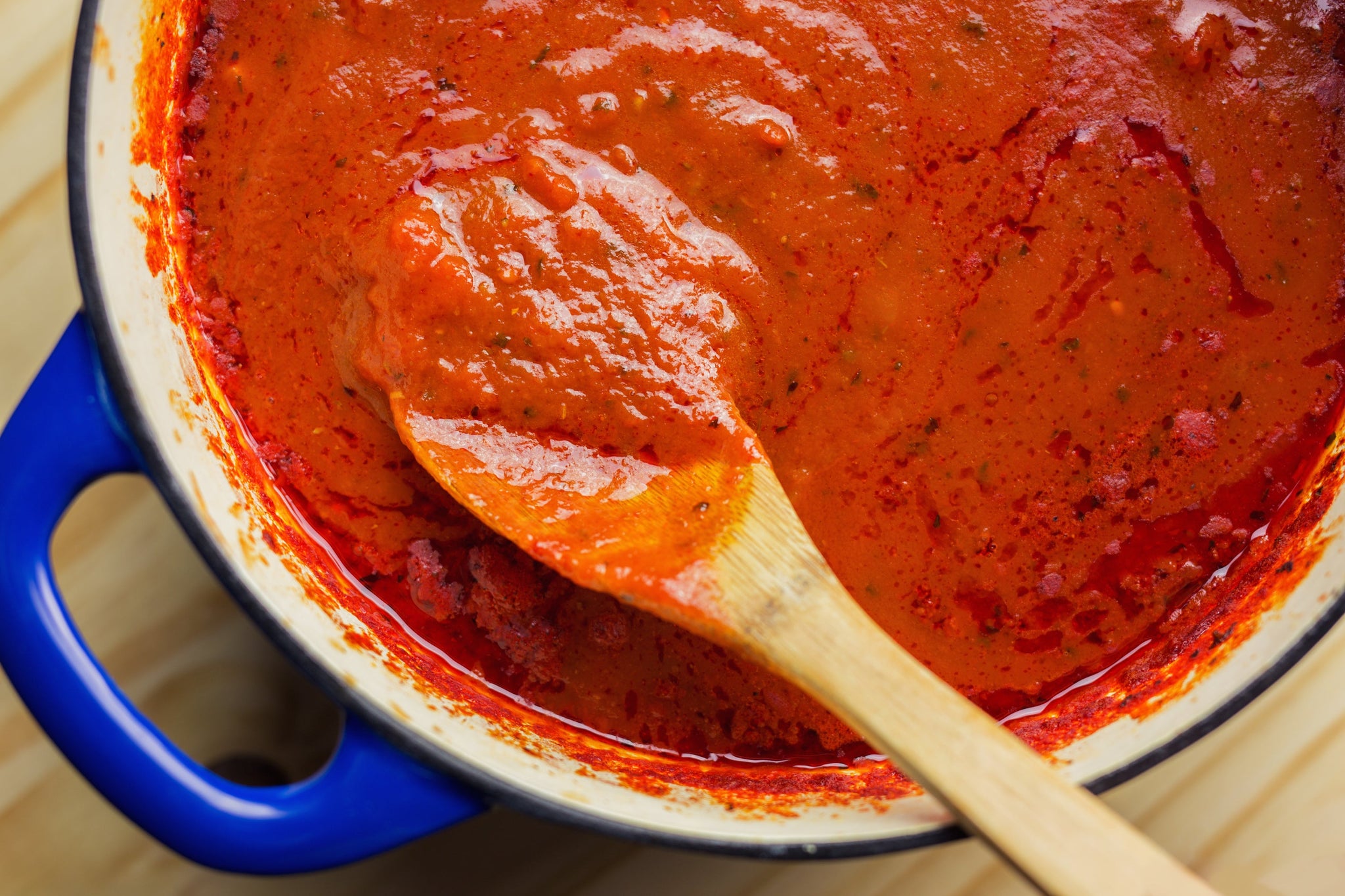 A wooden spoon rests in a pot of thick, red tomato sauce, showing a rich texture and vibrant color against the white and blue pot on a wooden surface.