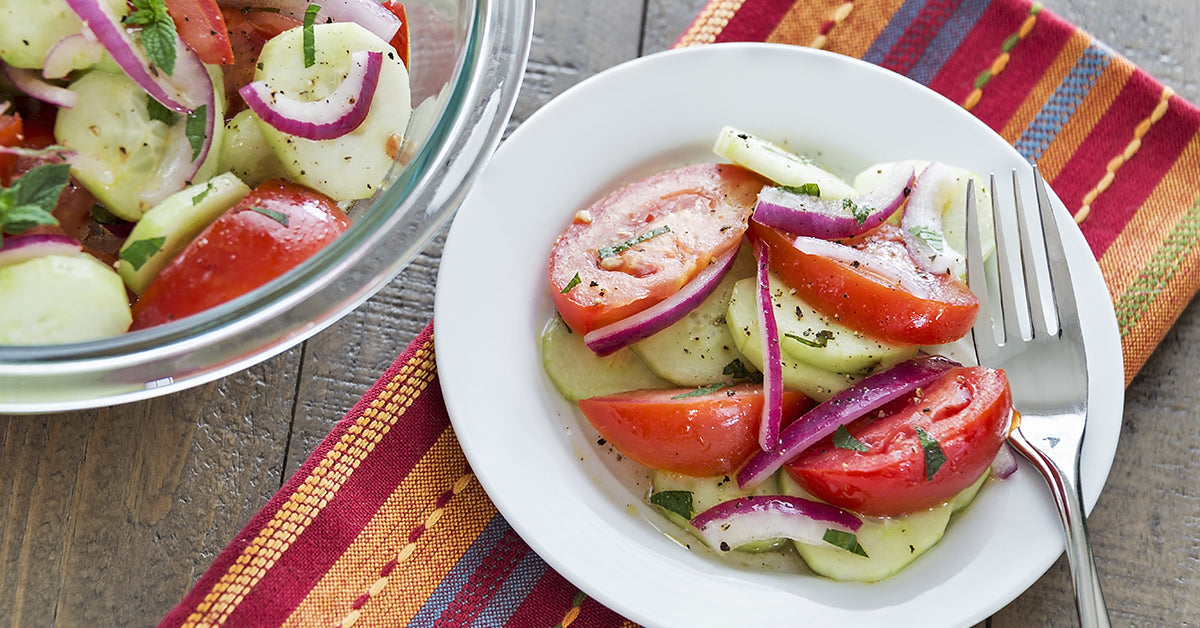 Plate of cucumber, tomato, and red onion salad with herbs beside a larger bowl of the same salad.