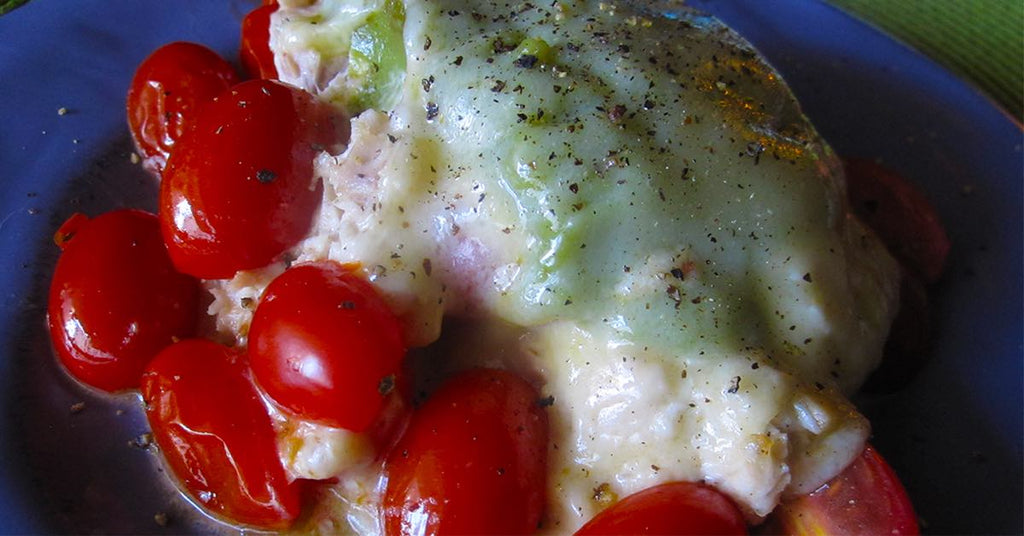A plate of baked chicken topped with melted cheese and green peppers, surrounded by bright red cherry tomatoes, garnished with black pepper.