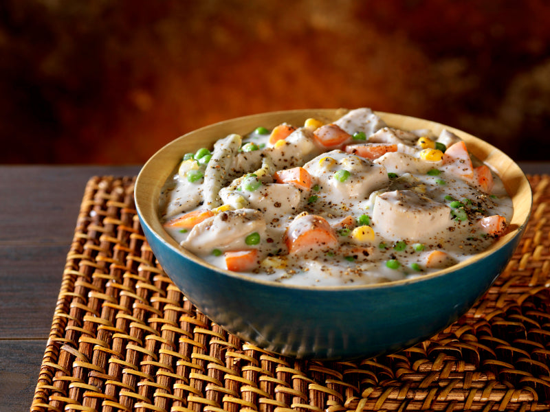 Creamy chicken stew with carrots, peas, and corn served in a blue bowl on a woven placemat.