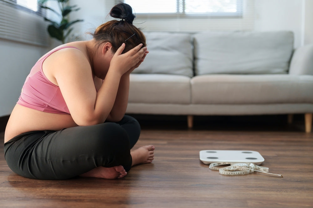 A person sits on the floor with their head in their hands, looking distressed, next to a scale and measuring tape in a bright, modern living room.