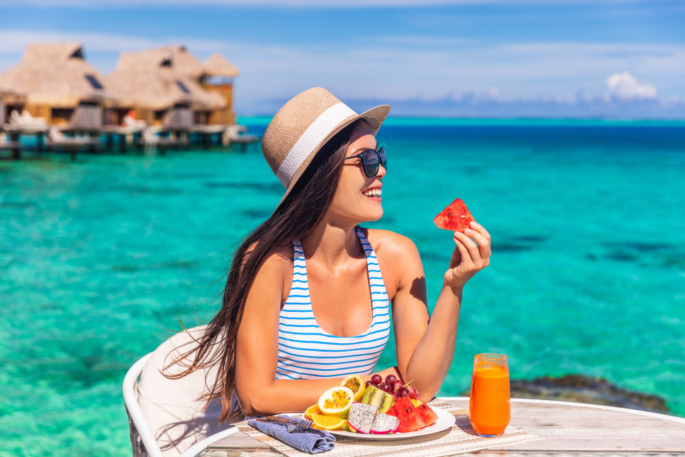 A woman in a striped swimsuit and sunhat smiles while enjoying a slice of watermelon at a table by the turquoise ocean, with a colorful fruit plate and a glass of orange juice in front of her.