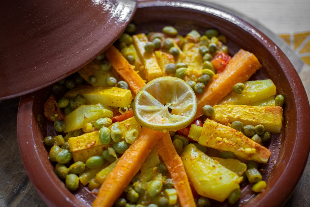 A clay tagine dish filled with colorful vegetables, including carrots, zucchini, and peas, topped with a slice of lemon, is partially covered by the lid, showcasing a vibrant, flavorful meal.