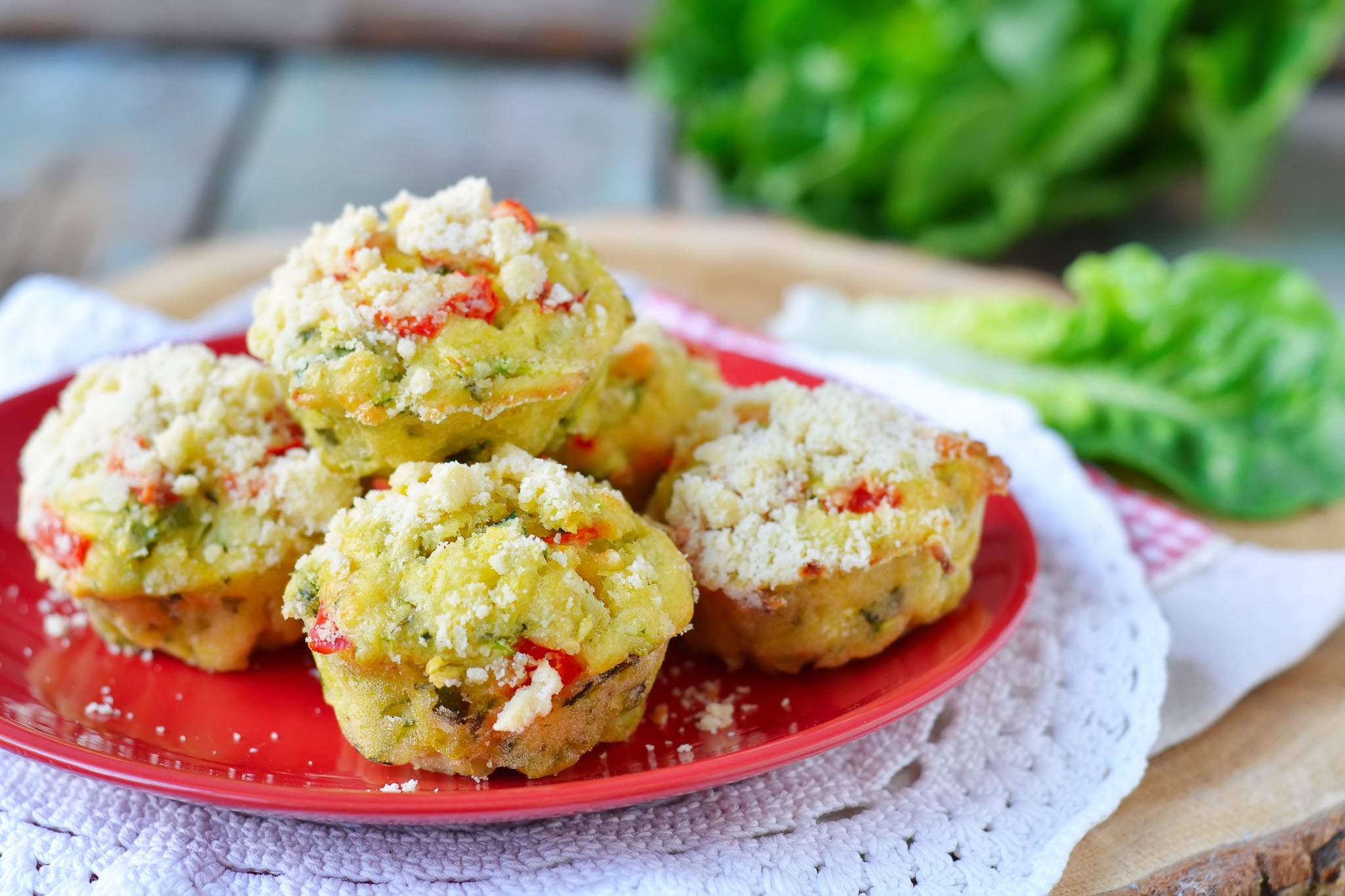 Four golden, baked vegetable muffins topped with crumbled cheese sit on a red plate, surrounded by a white lace doily, with fresh green lettuce in the background.