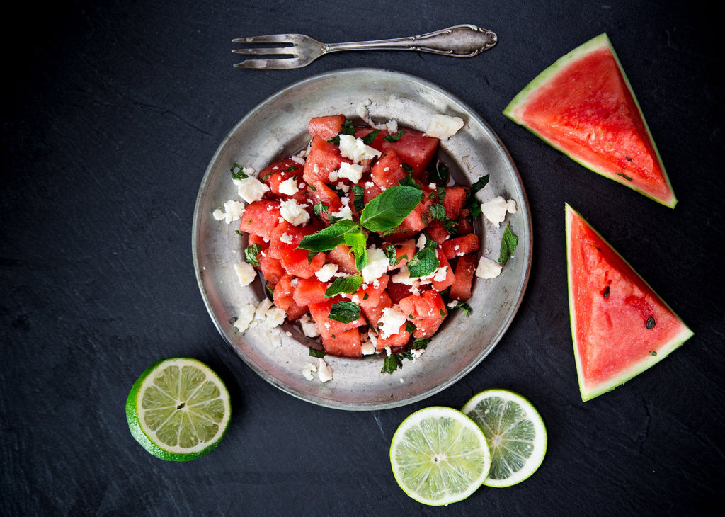 Bowl of watermelon salad with feta and mint, surrounded by lime slices and watermelon wedges on a dark surface