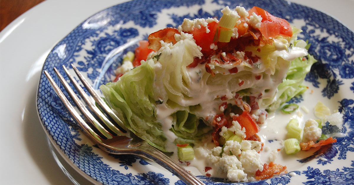 Iceberg lettuce wedge topped with creamy dressing, tomatoes, bacon, green onions, and blue cheese.