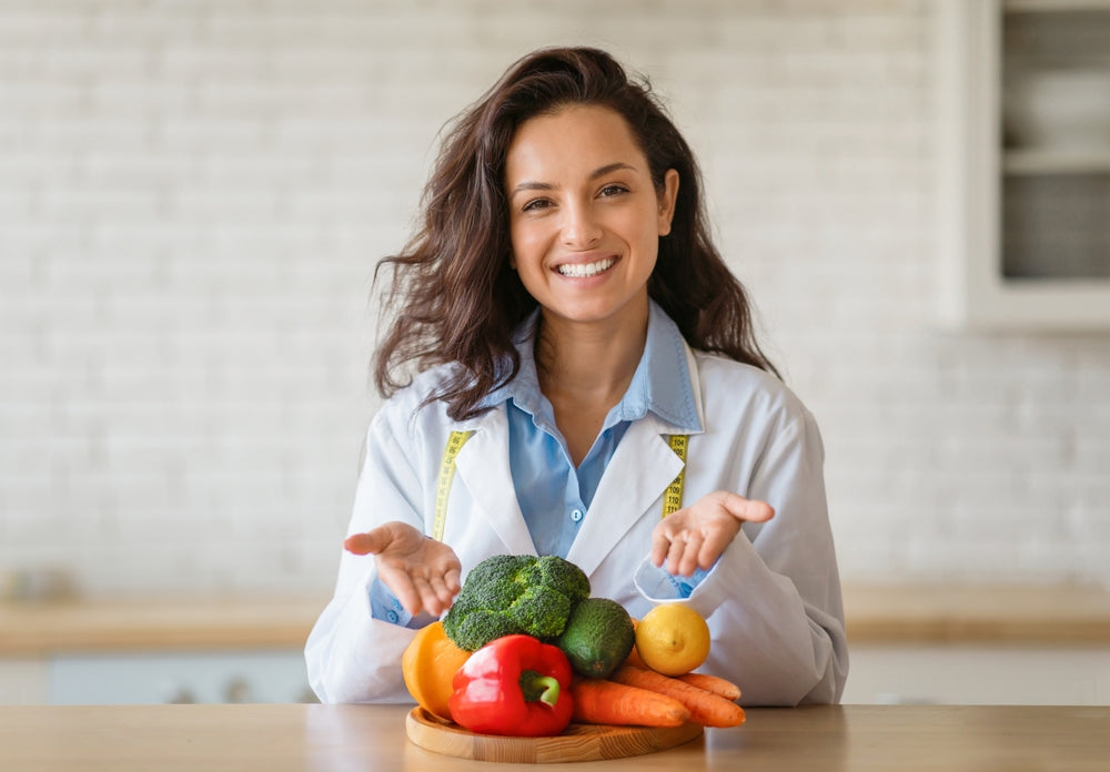 A smiling woman in a lab coat stands behind a wooden table, presenting a variety of fresh vegetables and fruits, including broccoli, peppers, and carrots, in a bright kitchen setting.