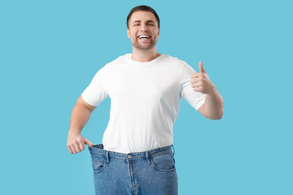 A smiling man in a white t-shirt gives a thumbs-up while holding up the waistband of oversized blue jeans against a light blue background, celebrating his weight loss success.