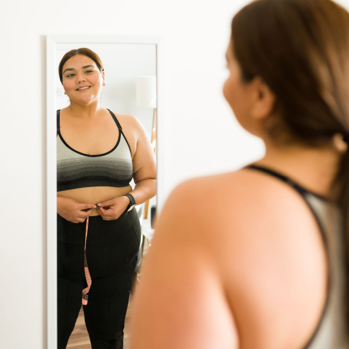 A woman in a sports bra smiles at her reflection in a mirror while holding a measuring tape around her waist, indicating a focus on her weight loss journey in a bright, modern room.