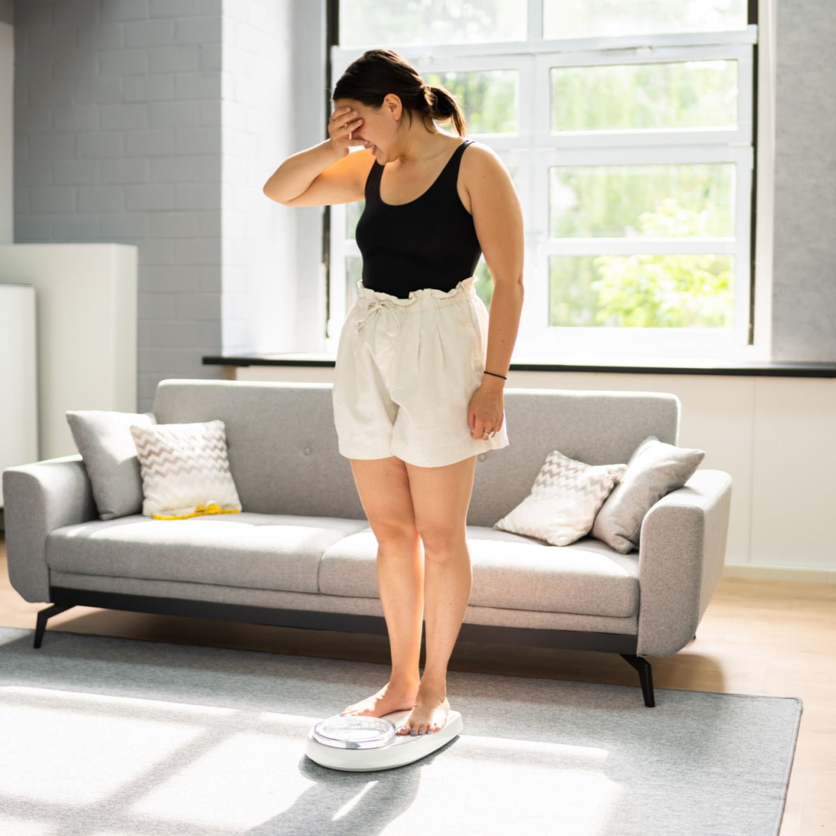 A woman stands barefoot on a scale in a bright living room, looking down with a worried expression while covering her face with one hand.