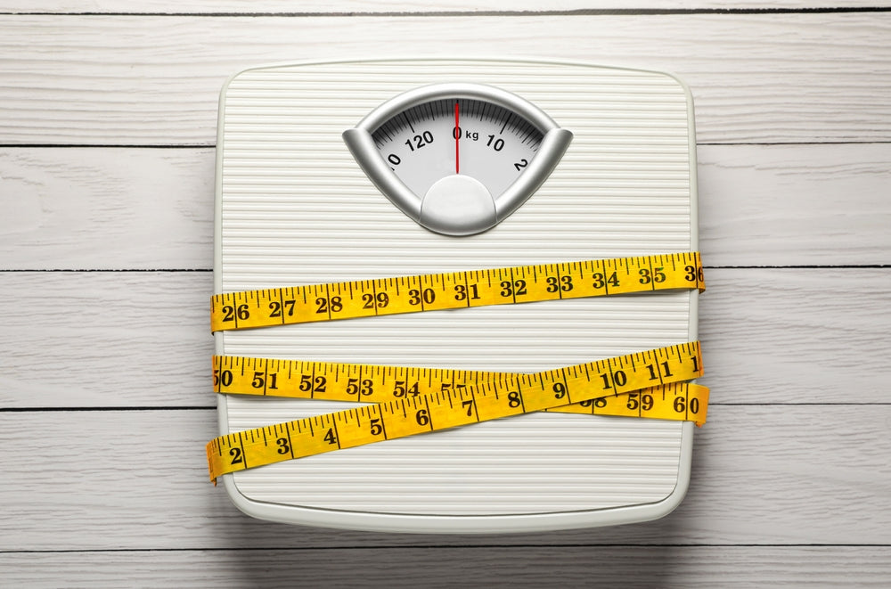 A white bathroom scale is placed on a wooden surface, with two yellow measuring tapes wrapped around it, symbolizing weight measurement and body size in the context of weight loss discussions.