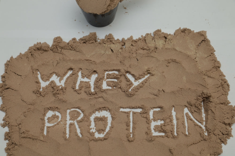 Brown whey protein powder is spread out on a surface, with the words "WHEY PROTEIN" written in white, while a small cup of the powder sits nearby.