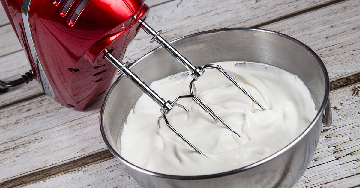 Hand mixer beating a bowl of thick, white whipped mixture on a wooden surface.