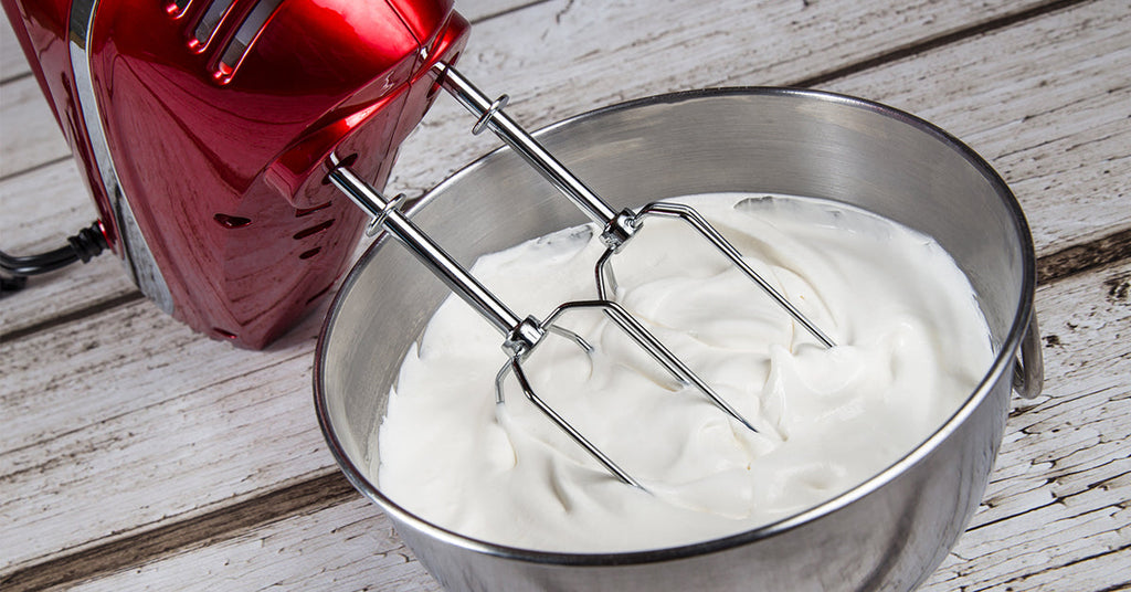 Hand mixer beating a bowl of thick, white whipped mixture on a wooden surface.