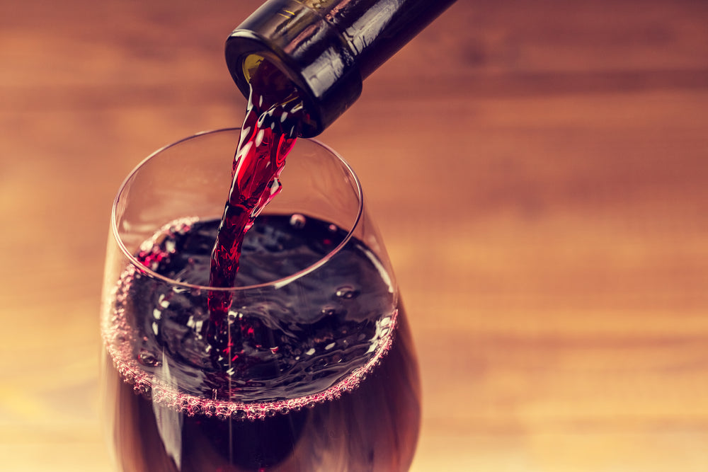 A bottle of red wine is being poured into a glass, creating ripples in the liquid, with a warm wooden surface in the background.