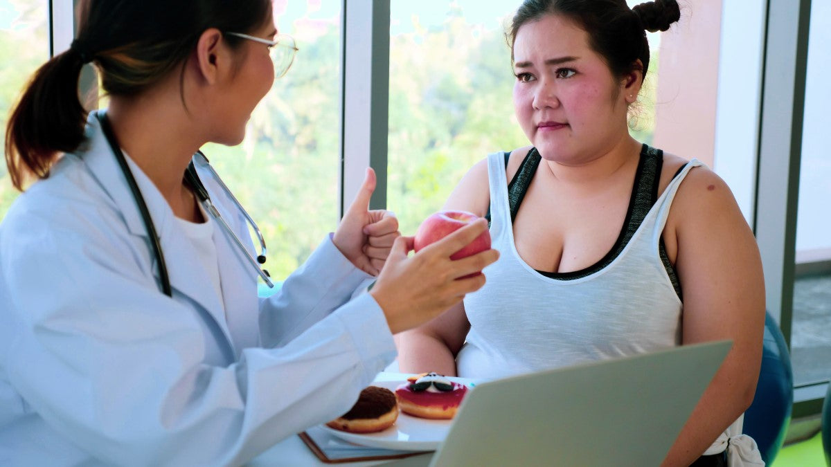 A doctor in a white coat holds an apple while discussing dietary choices with a concerned patient, who sits at a table with donuts, in a bright, modern office setting.