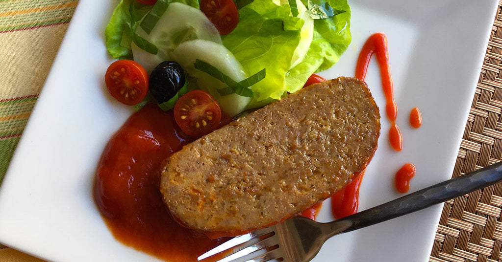 Slice of meatloaf with tomato sauce alongside lettuce, cucumbers, cherry tomatoes, and an olive.
