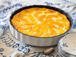 Peach tart in a round baking dish, topped with neatly arranged peach slices, set on a patterned tablecloth with plates nearby.