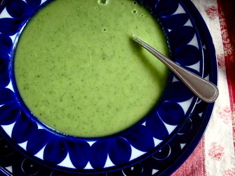 A bowl of smooth green soup sits on a decorative blue plate, with a silver spoon resting on the edge, against a textured red and white tablecloth background.