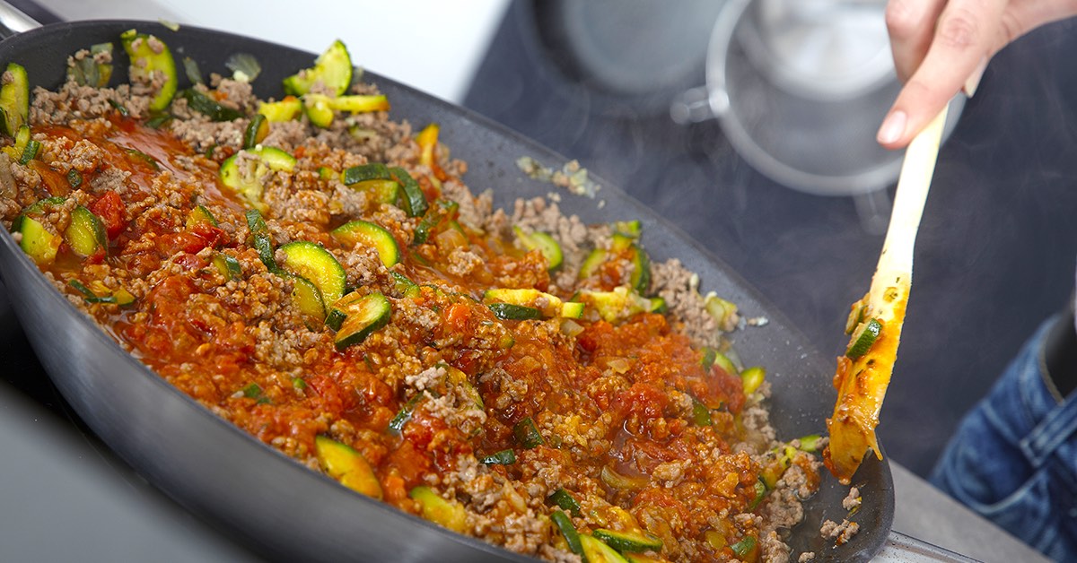 Person cooking a pan of minced meat, zucchini slices, and tomato sauce being stirred with a wooden spatula.