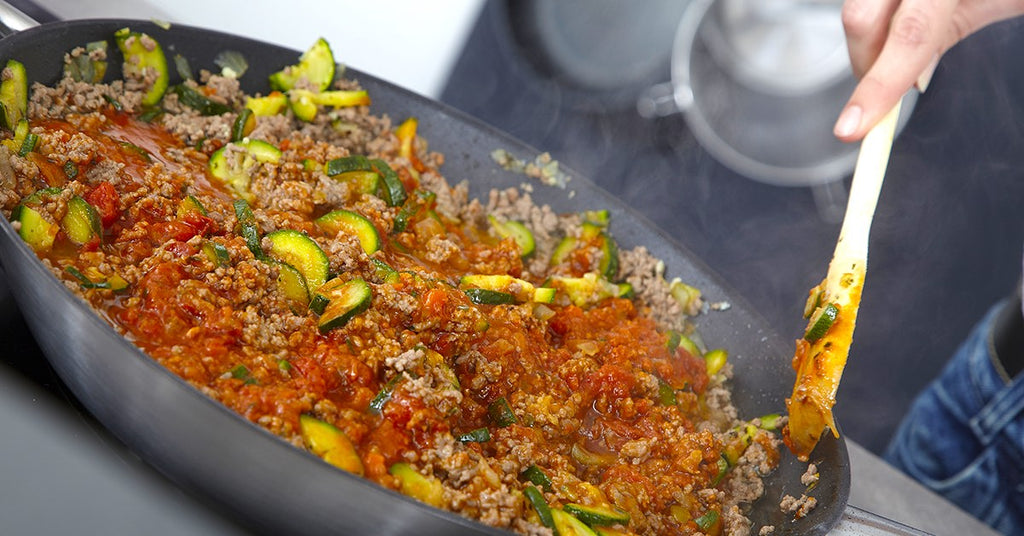 Person stirring a skillet of ground meat, zucchini, and tomato sauce simmering on the stove.