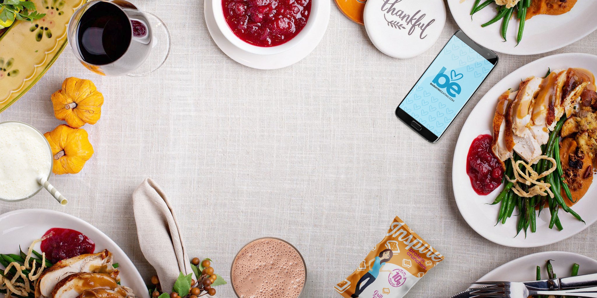 Thanksgiving-style table with turkey plates, cranberry sauce, drinks, mini pumpkins, and a phone on a beige tablecloth.